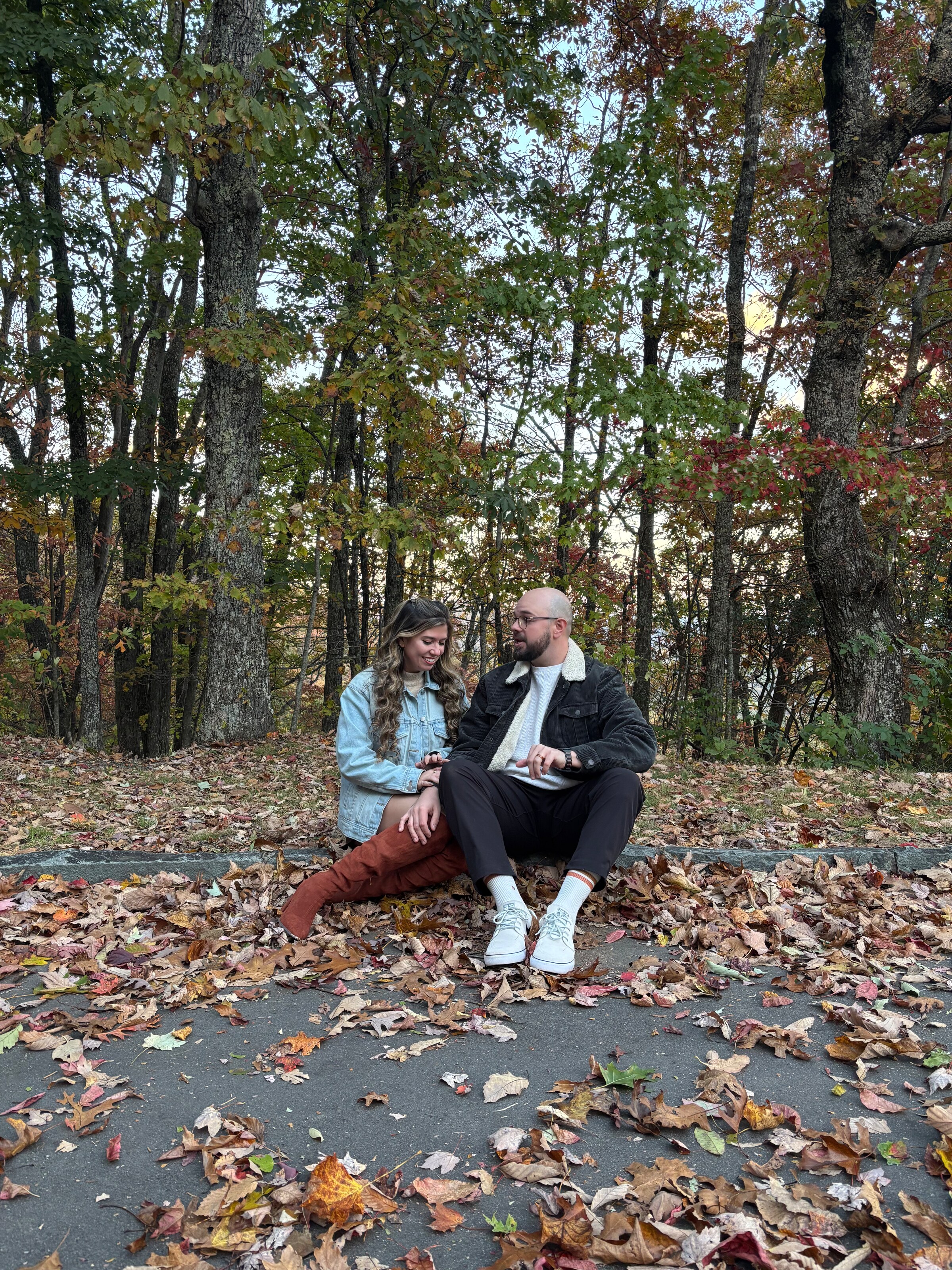 Sitting together on the edge of a path buried in fallen leaves, mid-conversation, bare autumn trees behind them.