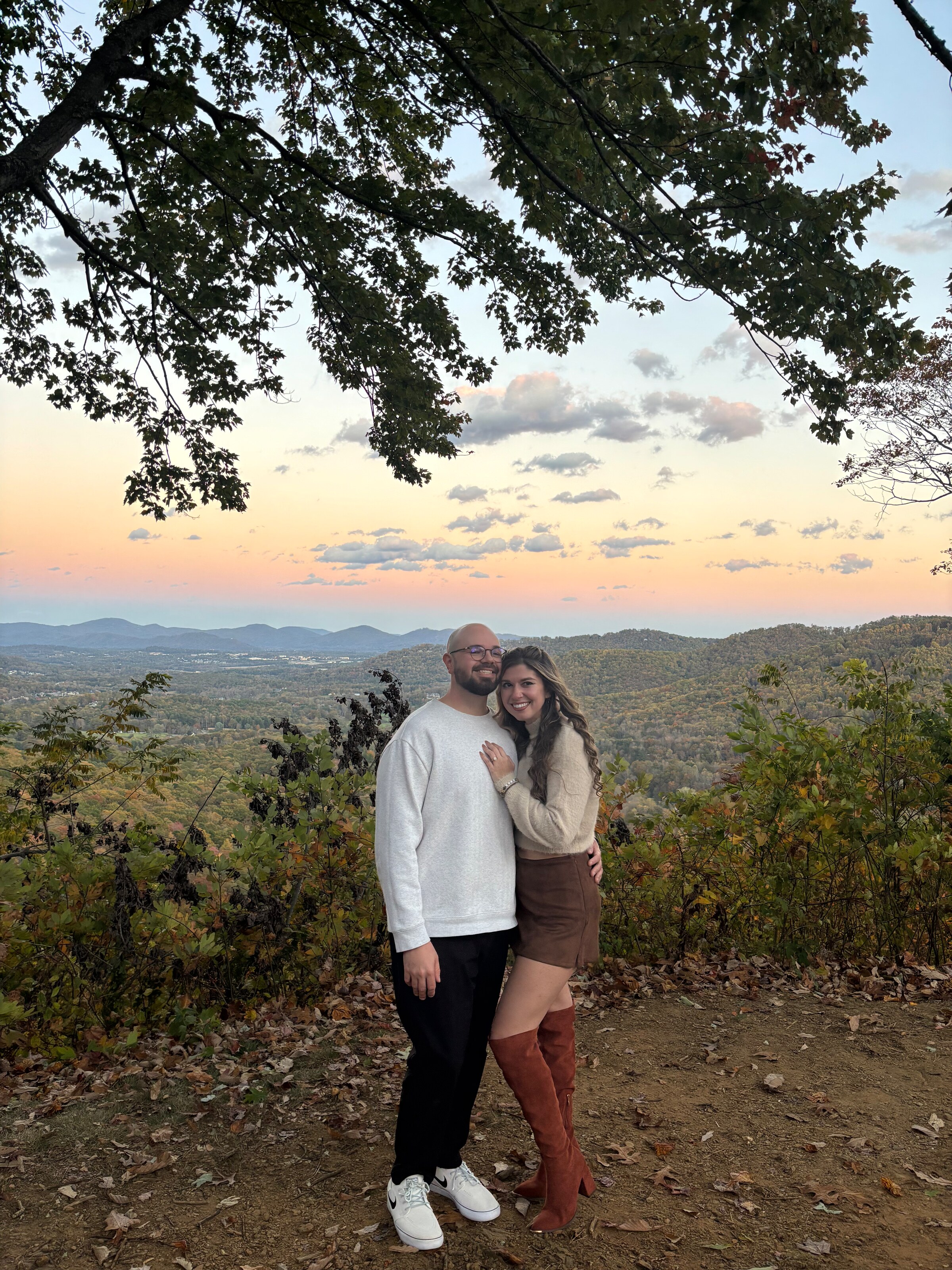 Nicole and Ian at a mountain overlook, arm in arm beneath a leafy branch, with a pale sunset over the ridges behind them.