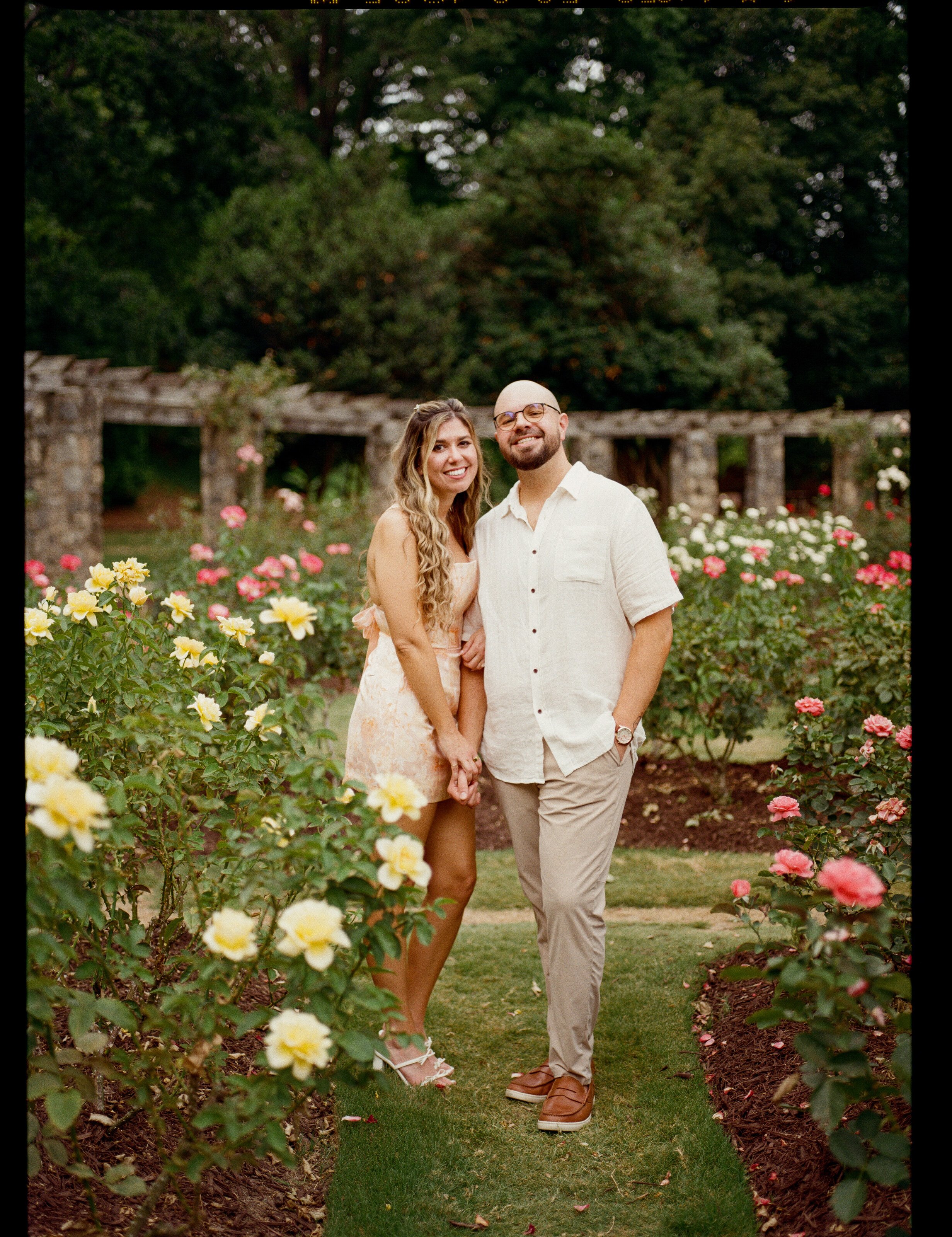 Standing together on the lawn between beds of yellow and pink roses, a long stone pergola behind them.