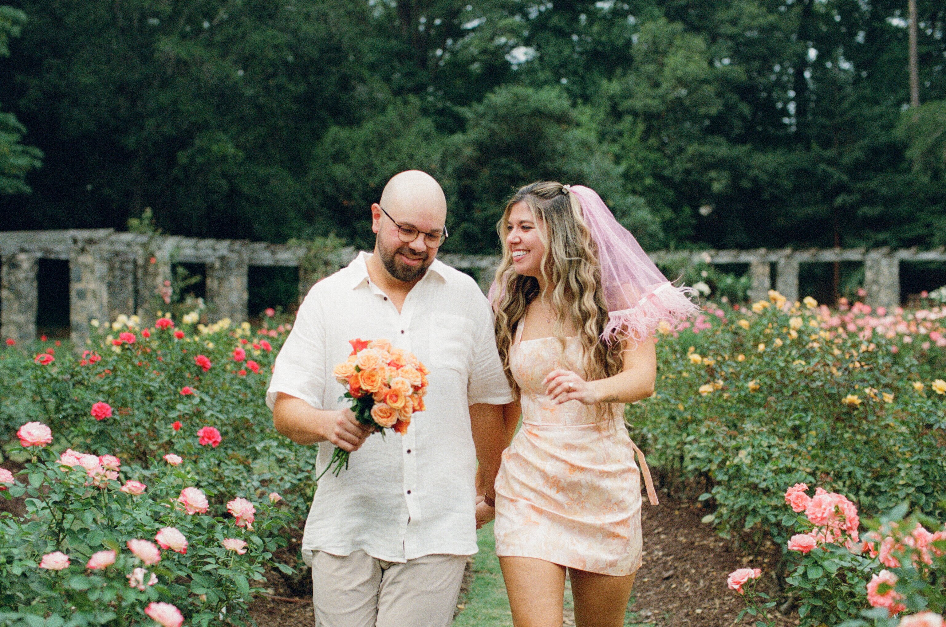 Walking hand in hand along a grass path through the rose beds, one carrying a bunch of orange roses, the other wearing a short pink veil.