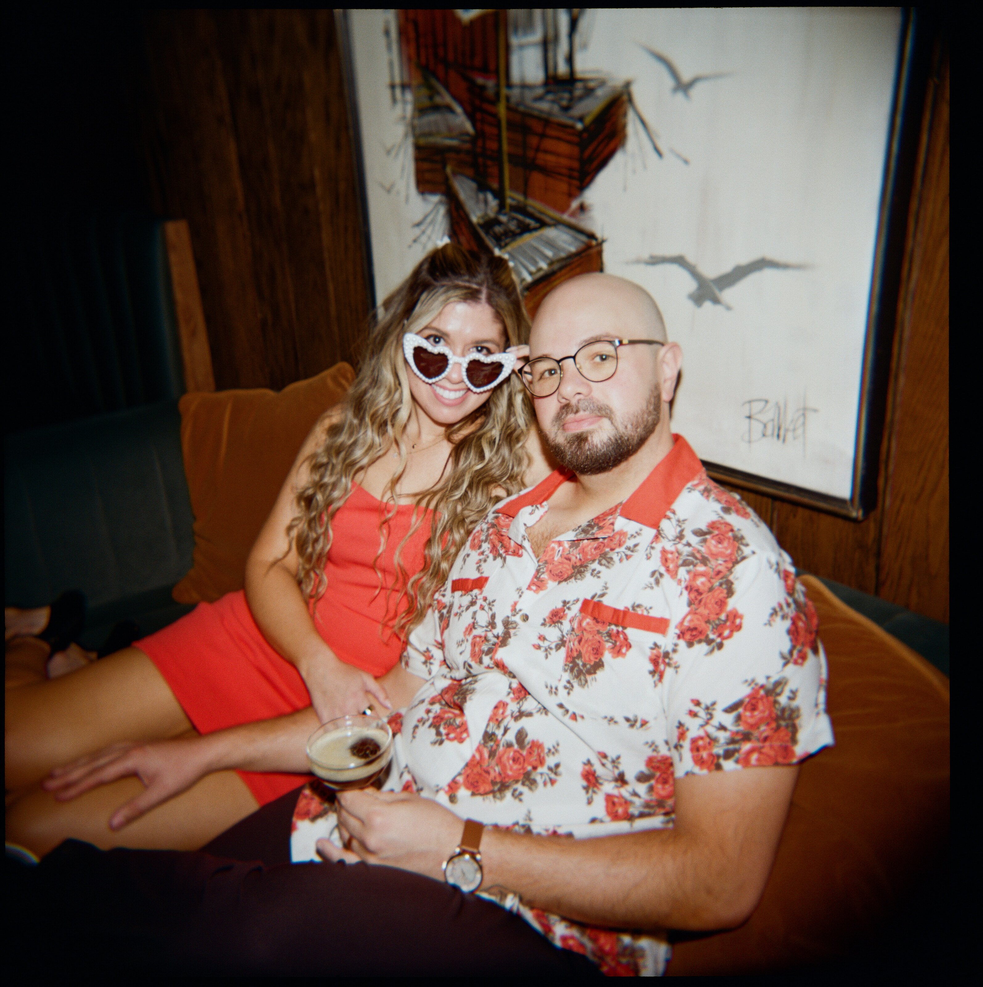 On a velvet couch beneath a painting, one wearing white heart-shaped sunglasses, the other holding a drink.