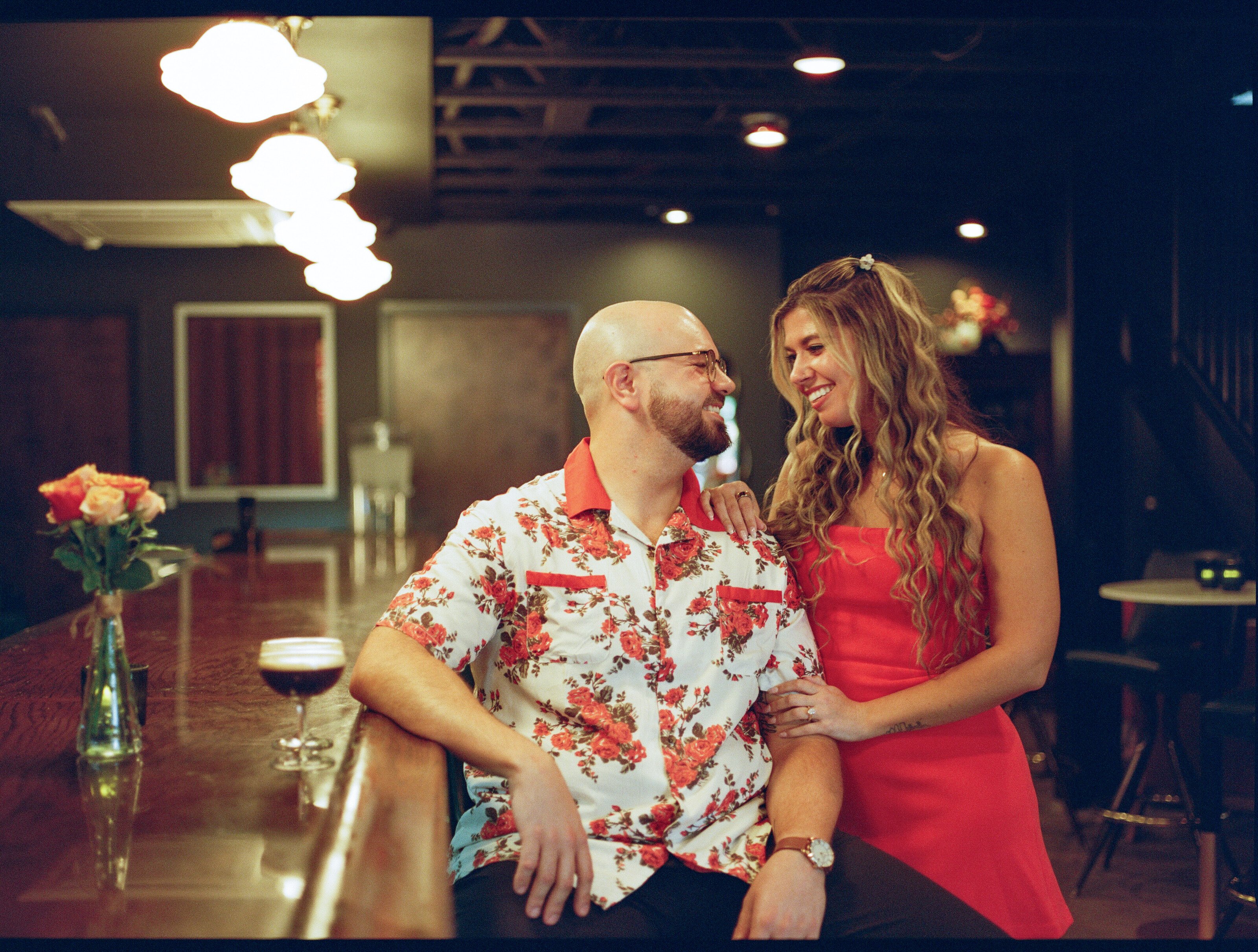 Leaning against a bar counter, laughing at each other, an espresso martini and a vase of roses beside them.