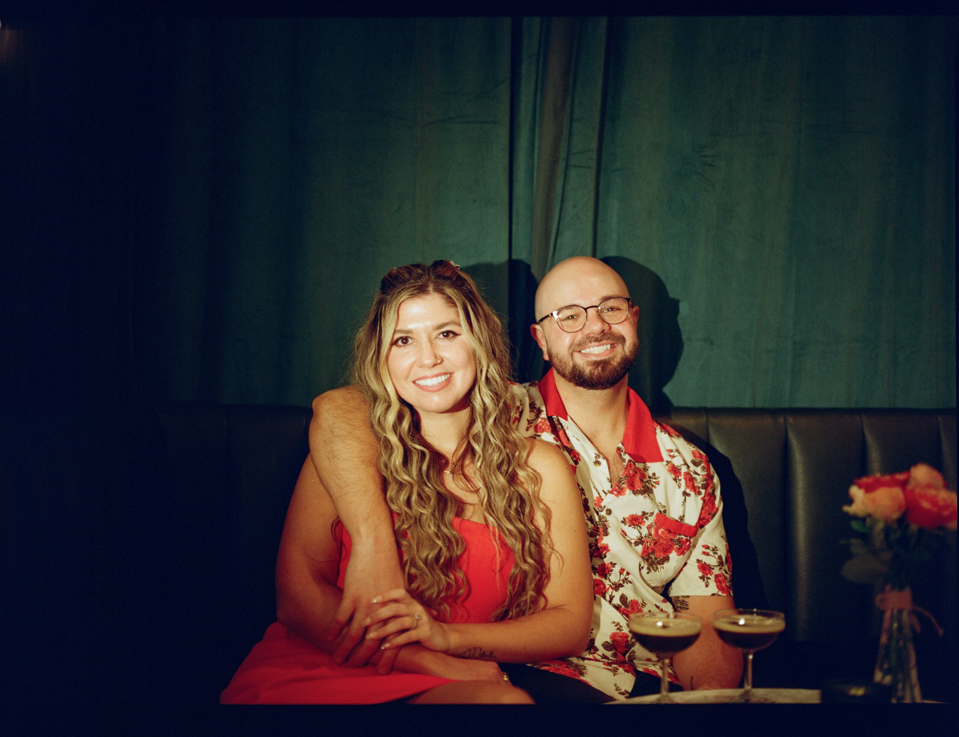 Side by side in a booth against a deep green curtain, two espresso martinis and a vase of roses on the table.