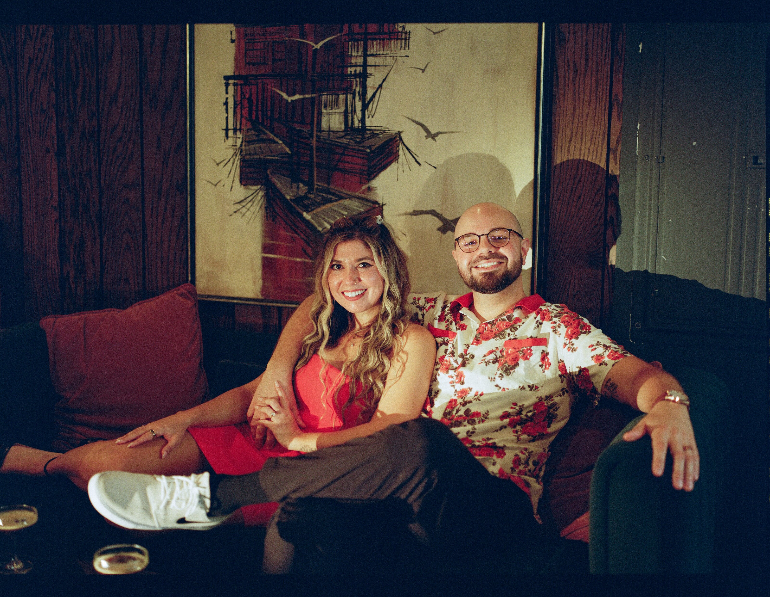 Settled into a red couch under a large painting, feet up, grinning at the camera.